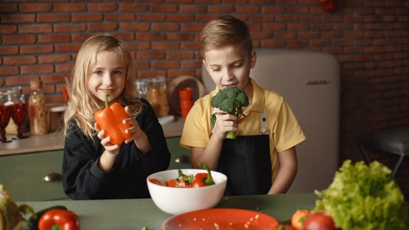 Cute girl and boy with fresh pepper and broccoli in hands standing near desk with assorted vegetables and salad bowl during vegetarian meal preparation