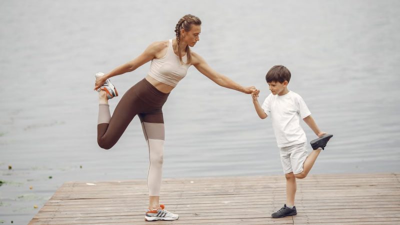 A mother and son holding a yoga pose together on a wooden pier by the water, promoting family fitness.