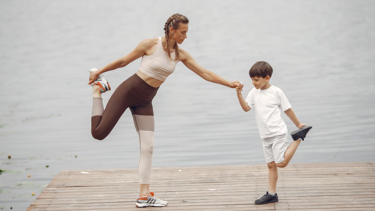 A mother and son holding a yoga pose together on a wooden pier by the water, promoting family fitness.